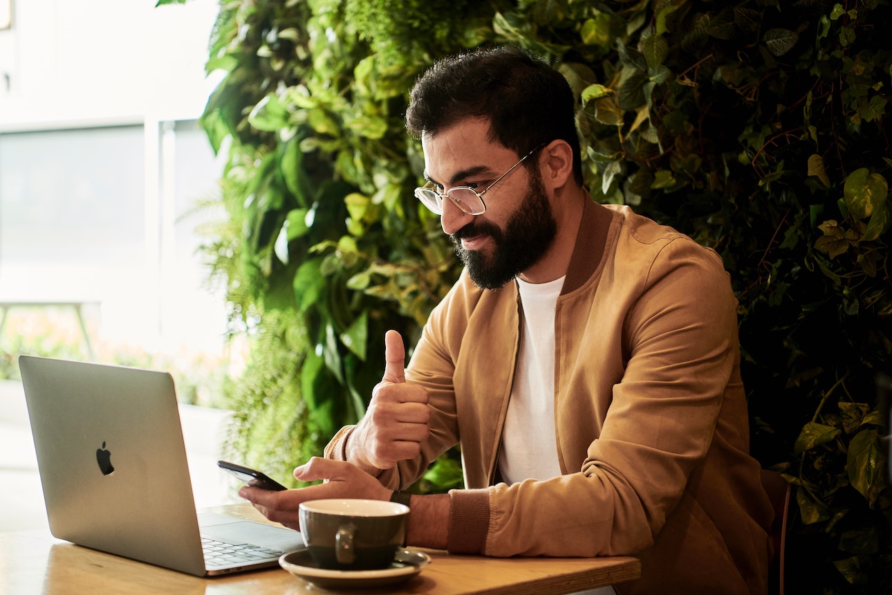 Man with beard holding a thumbs up in front of laptop and mobile phone, having a coffee