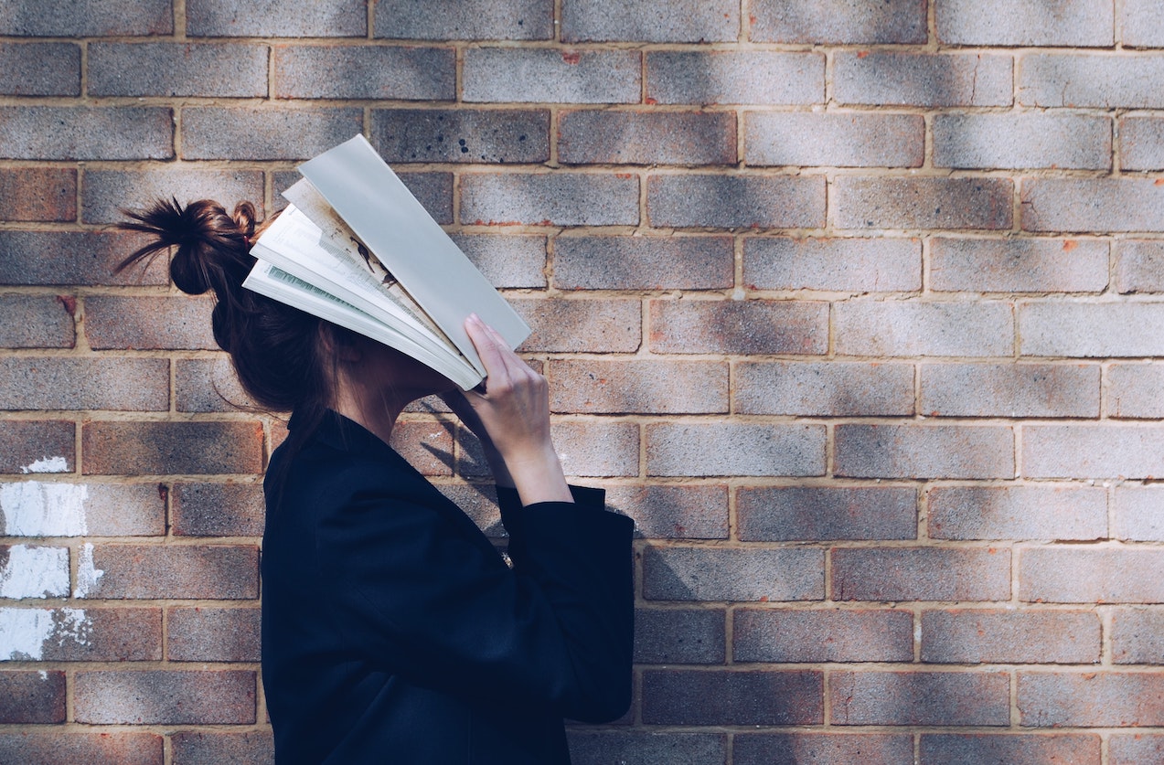 Writing challenges - Woman in black jacket in front of brick wall with a book covering her face