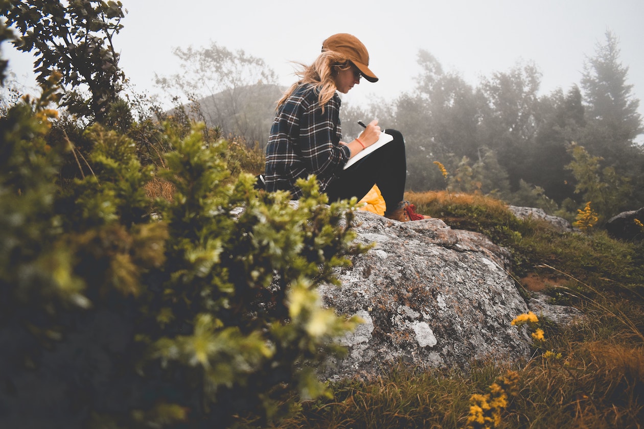 A woman in checkered shirt and cap writing in notebook on a hill