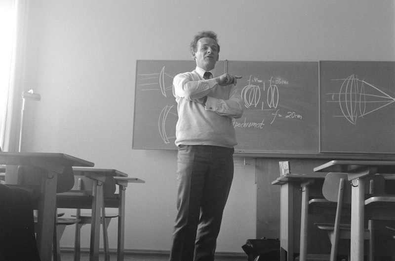 An old school pic of a teacher in sweater and tie standing in front of a blackboard