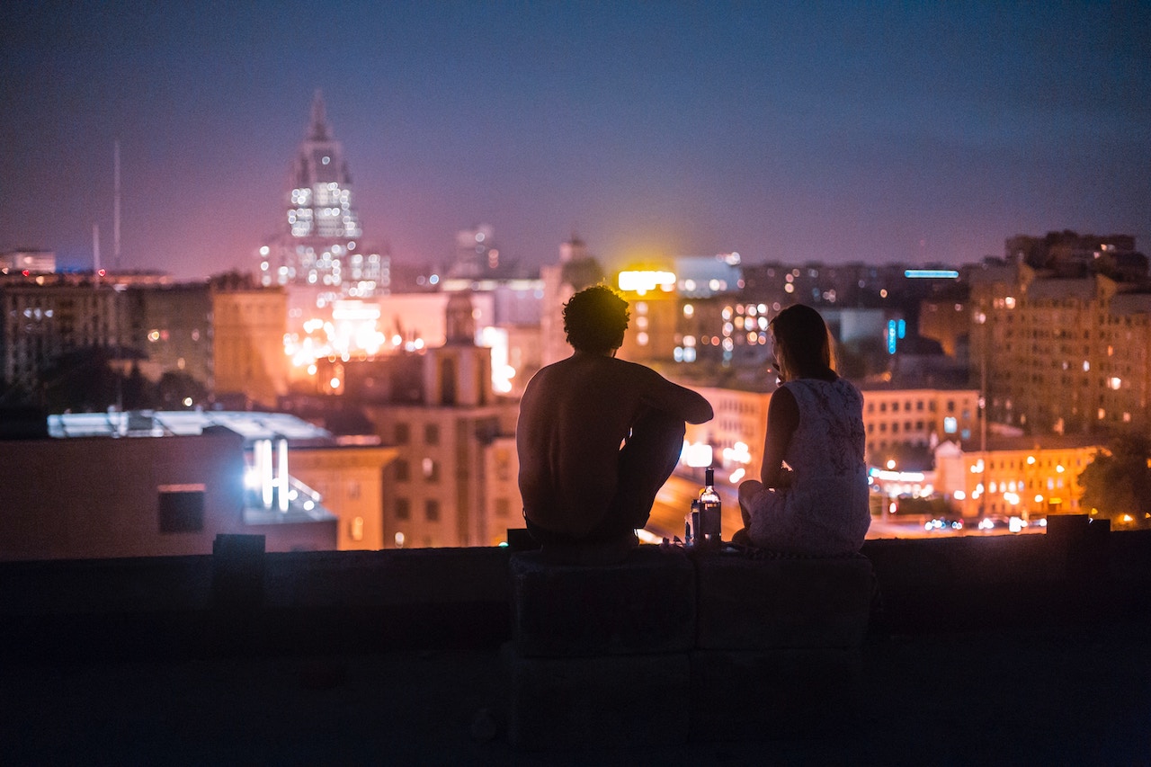 A couple sitting on the uppermost floor of apartments with bottle of whiskey between them, overlooking night cityscape
